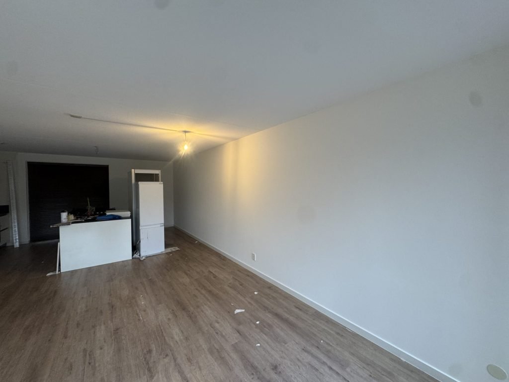Woonkamer met laminaatvloer en strakke witte wanden, met de keuken op de achtergrond